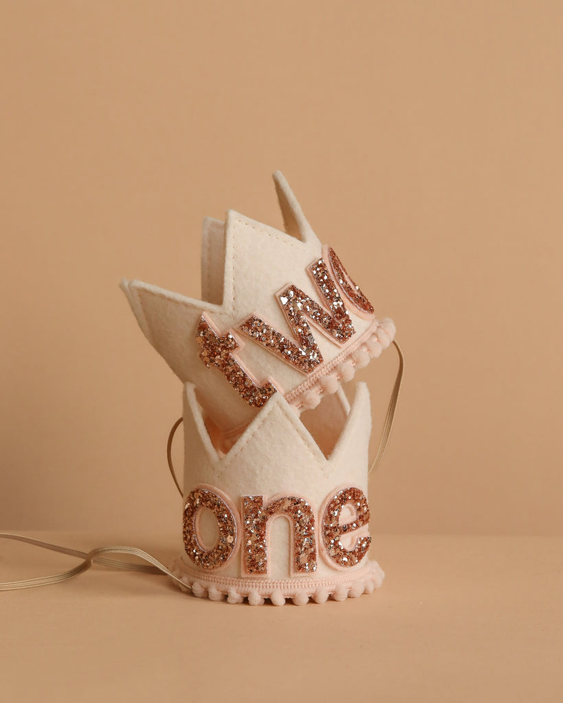 Two small Linen and Rose Crowns, each in cream felt with gold glittery "one" and "two" letters. Perfect party hats for birthdays or photo shoots; feature decorative edges, ribbon ties, and are displayed on a beige surface.