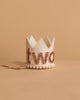 The Linen and Rose Crown is a small white felt crown with "two" in rose gold glitter, ideal for birthdays. It rests on a neutral beige surface and features an attached ribbon for easy wearing.