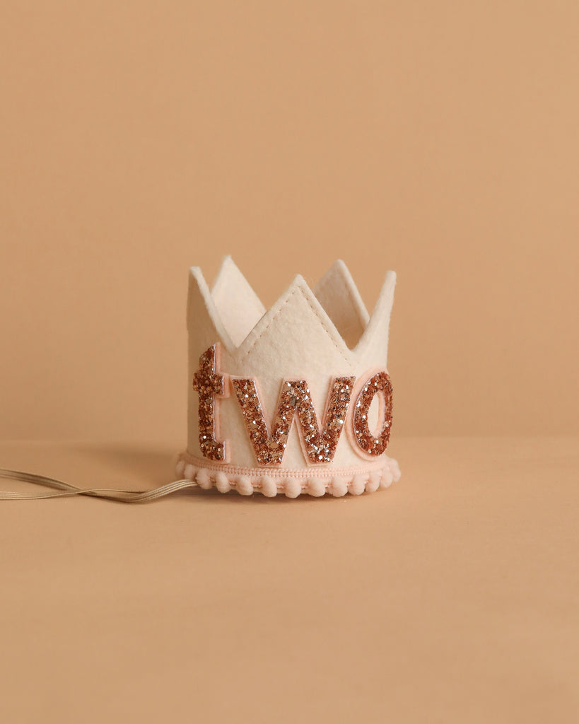 The Linen and Rose Crown is a small white felt crown with "two" in rose gold glitter, ideal for birthdays. It rests on a neutral beige surface and features an attached ribbon for easy wearing.