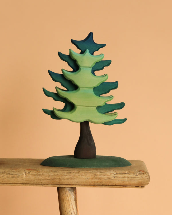 The Bumbu Handmade Wooden Extra Large Green Spruce Tree, featuring a stylized design with green and blue hues and a dark trunk, is displayed on a rustic wooden plank against a plain beige background, highlighting its handcrafted charm.