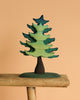 The Bumbu Handmade Wooden Extra Large Green Spruce Tree, featuring a stylized design with green and blue hues and a dark trunk, is displayed on a rustic wooden plank against a plain beige background, highlighting its handcrafted charm.