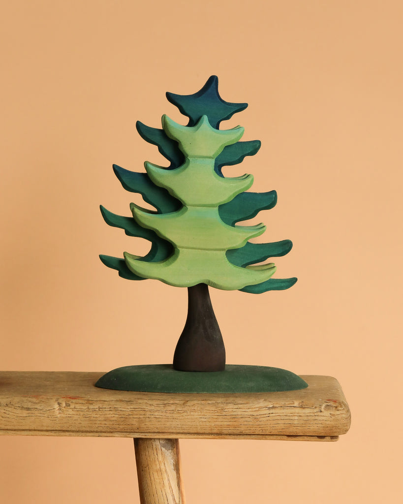 The Bumbu Handmade Wooden Extra Large Green Spruce Tree, featuring a stylized design with green and blue hues and a dark trunk, is displayed on a rustic wooden plank against a plain beige background, highlighting its handcrafted charm.