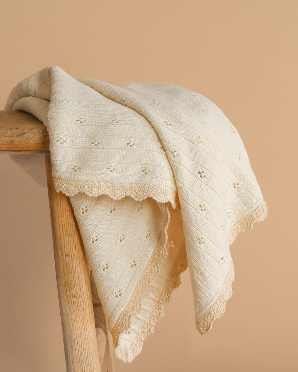 The Organic Pointelle Cotton Knit Baby Blanket, cream-colored and made in Peru from organic cotton with eyelet patterns and scalloped edges, is draped over a wooden stool against a beige background.