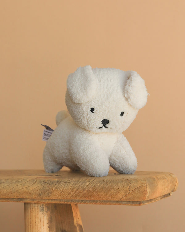 The Miffy Snuffy Stuffed Animal, a small white plush dog with floppy ears and black stitched features, sits on a rustic wooden stool against a beige background. Made by Bon Ton Toys with recycled PET filling.