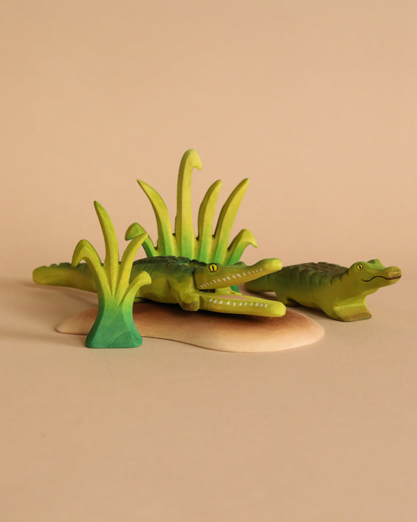 Two small green wooden crocodile figurines from the Bumbu Wooden Crocodile Cove Set rest on a beige surface with painted grass pieces, evoking a riverbank scene. Crafted from sustainably sourced wood, the background is plain and neutral.
