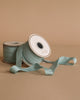 Two spools of Maileg Ribbon - Dusty Mint (10m), one upright and one lying down, are displayed on a neutral beige background. A length unrolls in front, ideal for adding an exclusive touch to decor or gift wrapping.