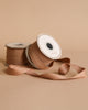 Two spools of Maileg Ribbon - Soft Brown (10m) rest on a beige background—one upright, one on its side with the ribbon unrolled—ideal for adding an exclusive touch to gift wrapping or special projects.