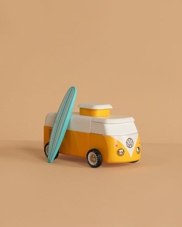 The VW Beach Bus Sunset is a small yellow and white Volkswagen Type 2 toy van with a surfboard leaning against its side, shown on a plain beige background.