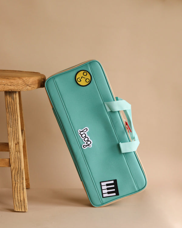 The Loog Piano Carrying Case, a teal water-resistant bag featuring patches of a yellow smiley face, "loog," and piano keys, is shown leaning against a wooden stool on a beige background.