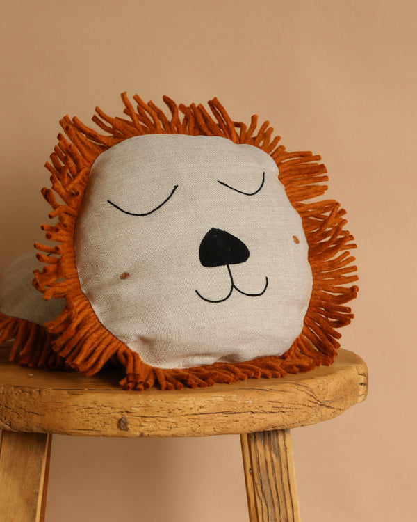 The Ferm Living Safari Cushion - Lion, with its soft linen, hand-embroidered details, closed eyes, and orange yarn mane, sits on a rustic wooden stool against a beige background—an adorable addition to any kids' room.