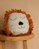 The Ferm Living Safari Cushion - Lion, with its soft linen, hand-embroidered details, closed eyes, and orange yarn mane, sits on a rustic wooden stool against a beige background—an adorable addition to any kids' room.
