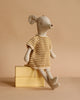 The Maileg Medium Mouse With Knitted Dress, dressed in yellow and white stripes, sits atop two stacked yellow books against a beige background.