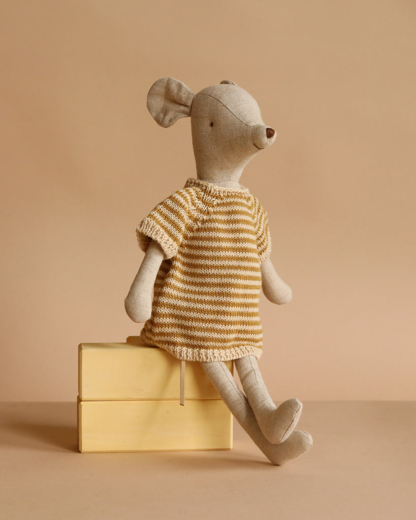 The Maileg Medium Mouse With Knitted Dress, dressed in yellow and white stripes, sits atop two stacked yellow books against a beige background.
