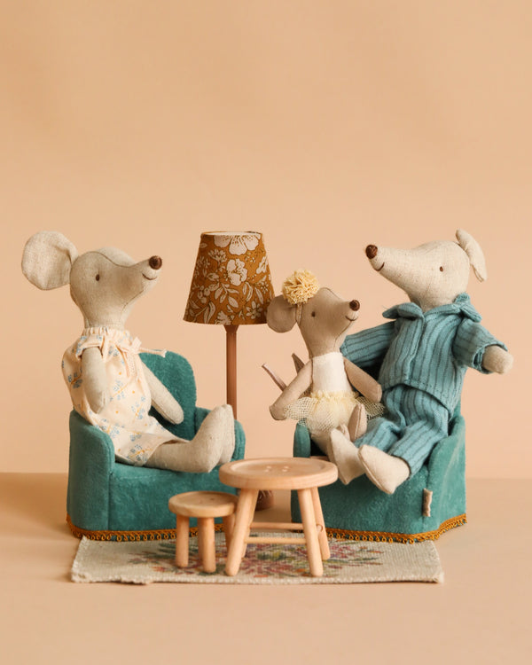 Three Maileg Mice dolls in pastel outfits relax on teal chairs around a small wooden table from the Maileg Living Room Set, complete with a vintage-style floor lamp and rug, creating a cozy scene against a beige background.