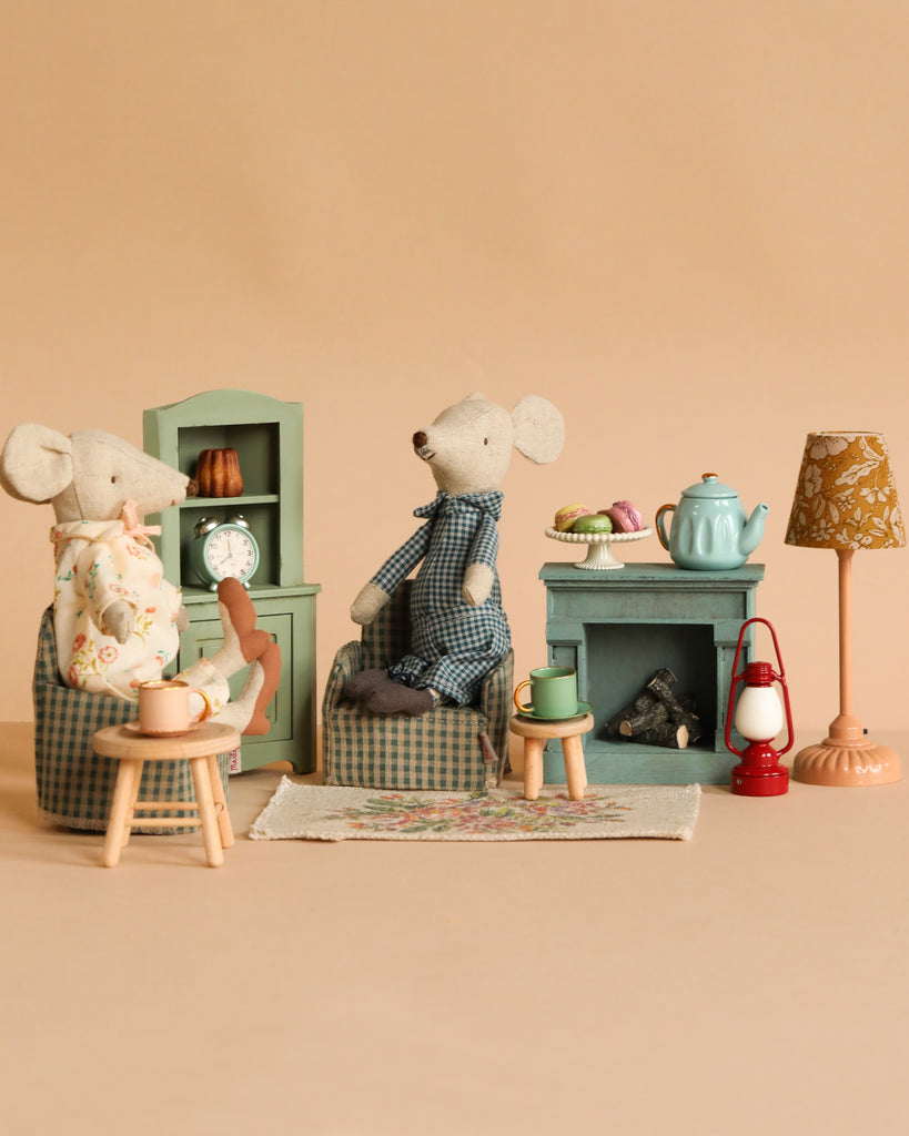The Maileg Grandma And Grandpa Afternoon Tea set features two mice relaxing in a cozy miniature living room with pastel furniture, a tea set, pastries, books, a lamp, and a clock—perfect for charming play on a beige backdrop.