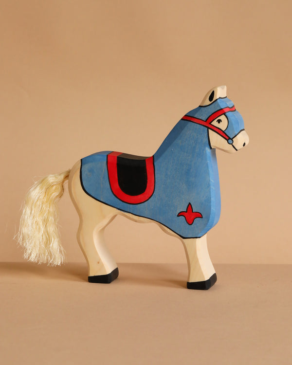 The Holztiger Tournament Horse, Blue is a handcrafted wood toy featuring a cream body, blue saddle and headpiece, red and black accents, and a stringy tail. Inspired by HOLZTIGER figures, it rests on a plain light brown surface.