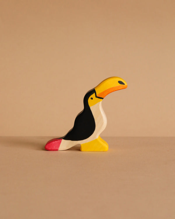 The Holztiger Toucan, a small handcrafted wooden figurine painted in black, white, yellow, orange, and pink, stands on a beige surface with a matching background. Ships in one week.