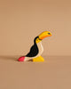 The Holztiger Toucan, a small handcrafted wooden figurine painted in black, white, yellow, orange, and pink, stands on a beige surface with a matching background. Ships in one week.