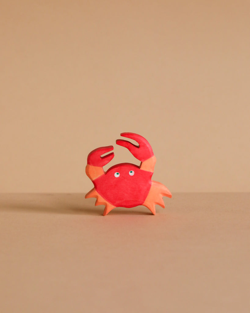 A small, red and orange cartoon crab with two raised claws and wide eyes stands on a light brown surface—just like the handcrafted Holztiger Crab, made in Europe and ships in one week.
