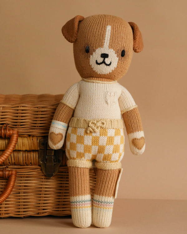 Cuddle + Kind George The Dog is a hand-knit doll in a cream shirt and yellow plaid shorts, standing upright before a wicker basket on a beige background. Filled with hypoallergenic polyfill for gentle cuddles.