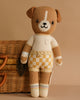 Cuddle + Kind George The Dog is a hand-knit doll in a cream shirt and yellow plaid shorts, standing upright before a wicker basket on a beige background. Filled with hypoallergenic polyfill for gentle cuddles.