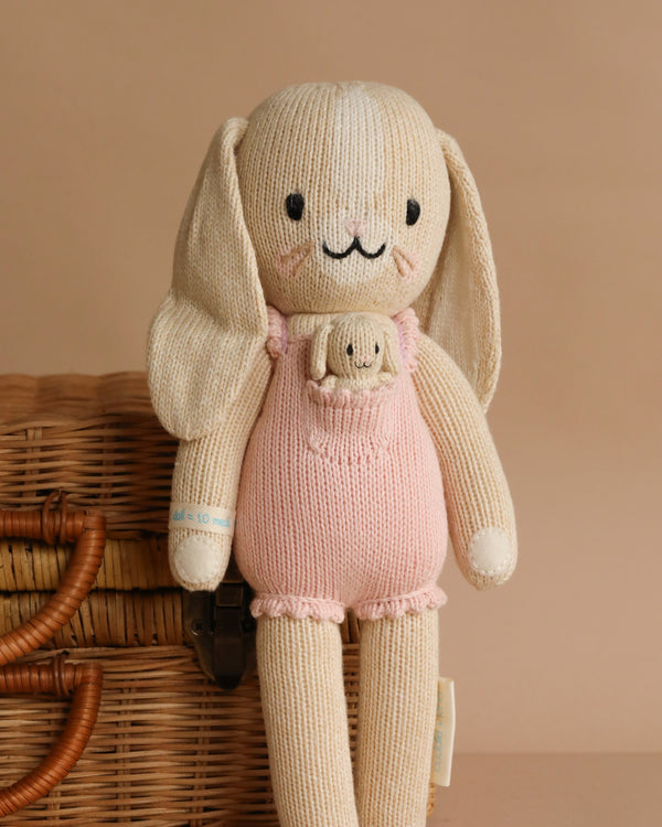Cuddle + Kind Briar The Bunny - Peony Pink is a handmade, hypoallergenic bunny doll in pink overalls with a tiny bunny in its pocket, perfect for gentle play and displayed before a wicker basket on a beige background.