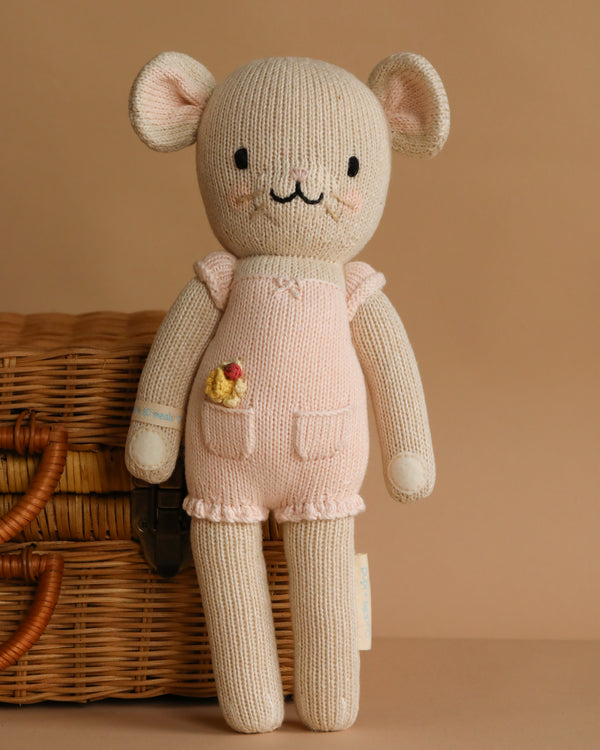 Cuddle + Kind Beatrice The Mouse - Blush is a handmade, knitted doll with a smiling face, light pink overalls, and a small yellow chick in the pocket, shown standing by a wicker basket against a beige background.