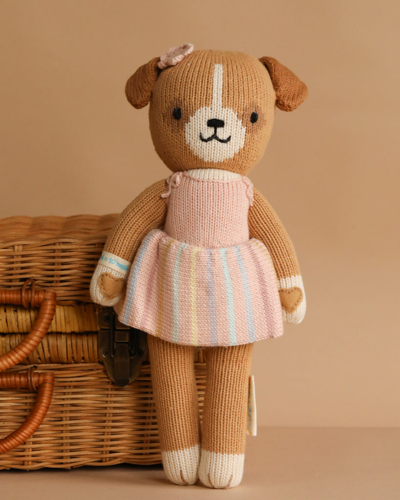 Cuddle + Kind Bea The Dog: a hand-knit brown stuffed dog in a pastel pink striped dress with a small ear bow, posed before a wicker picnic basket on a beige background.
