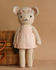 The Cuddle + Kind Emma The Mouse is a cream-colored, handmade knitted doll made from hypoallergenic materials, dressed in a light pink dress with a flower crown, shown standing before a wicker basket on a beige background.