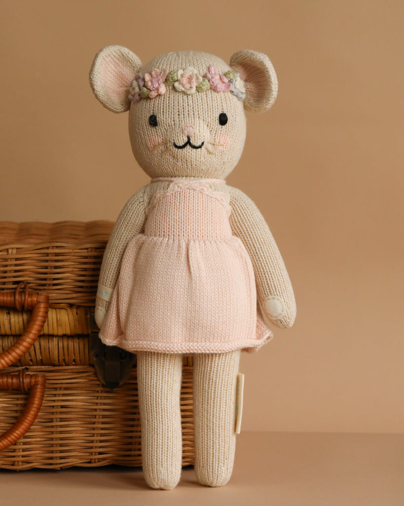The Cuddle + Kind Emma The Mouse is a cream-colored, handmade knitted doll made from hypoallergenic materials, dressed in a light pink dress with a flower crown, shown standing before a wicker basket on a beige background.