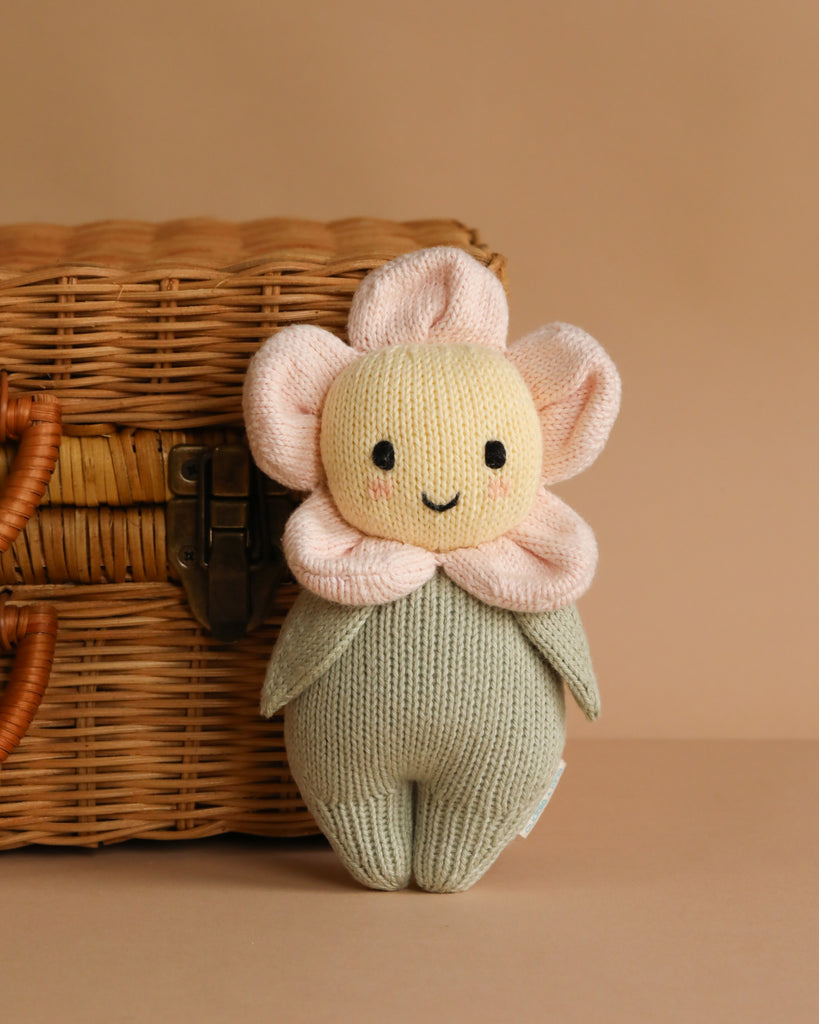 The Cuddle + Kind Baby Daisy - Blush Pink, a hand-knit plush toy with a smiling face, blush pink petals, and a green leaf body made of Peruvian cotton yarn, sits before a woven wicker basket on a beige background.