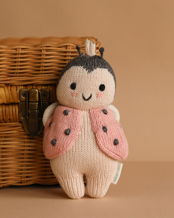 The Cuddle + Kind Baby Ladybug, a soft hand-knit doll made from natural Peruvian cotton yarn with a pink and black vest, smiling face, and antennae, sits upright near a wicker basket on a beige surface.