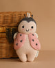 The Cuddle + Kind Baby Ladybug, a soft hand-knit doll made from natural Peruvian cotton yarn with a pink and black vest, smiling face, and antennae, sits upright near a wicker basket on a beige surface.