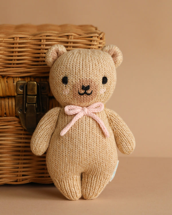 The Cuddle + Kind Baby Honey Bear - Blush, hand-knit from soft Peruvian cotton yarn in beige with a pink bow, stands before a woven wicker basket with a metal latch against a light brown background.