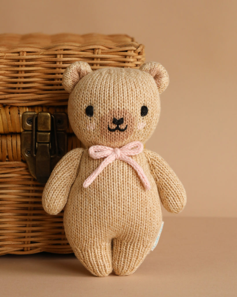 The Cuddle + Kind Baby Honey Bear - Blush, hand-knit from soft Peruvian cotton yarn in beige with a pink bow, stands before a woven wicker basket with a metal latch against a light brown background.