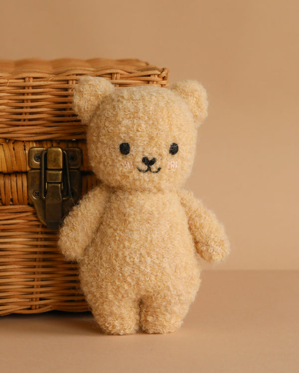 The Cuddle + Kind Baby Bouclé Bear - Honey, a small beige teddy with stitched eyes and a smiling mouth, stands in front of a wicker basket with a metal latch on a beige background.