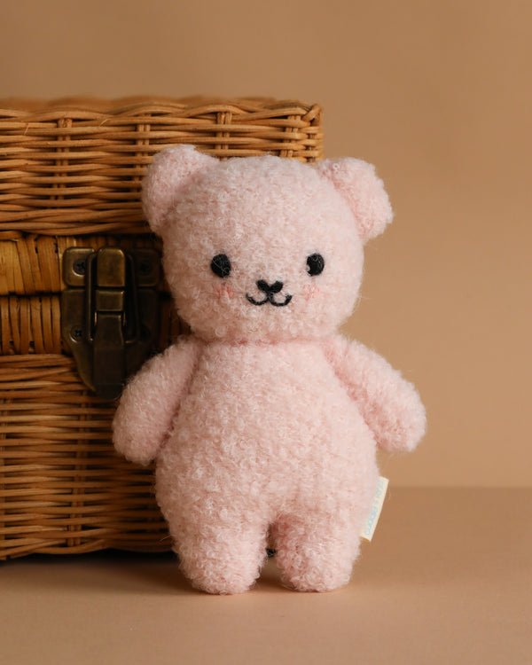 The Cuddle + Kind Baby Bouclé Bear - Strawberry, a soft, pink bear with a smiling face, stands in front of a brown wicker basket against a beige background.