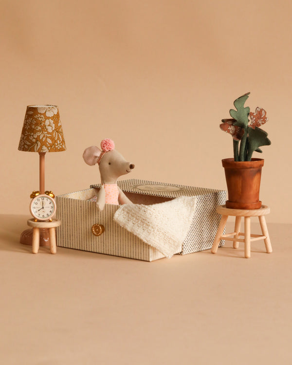 A Maileg Dancing Mouse in Daybed – Cozy Morning Set rests in a tiny bed, while a table and stool nearby display a lamp, clock, and plant against a beige background.