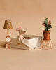 A Maileg Dancing Mouse in Daybed – Cozy Morning Set rests in a tiny bed, while a table and stool nearby display a lamp, clock, and plant against a beige background.