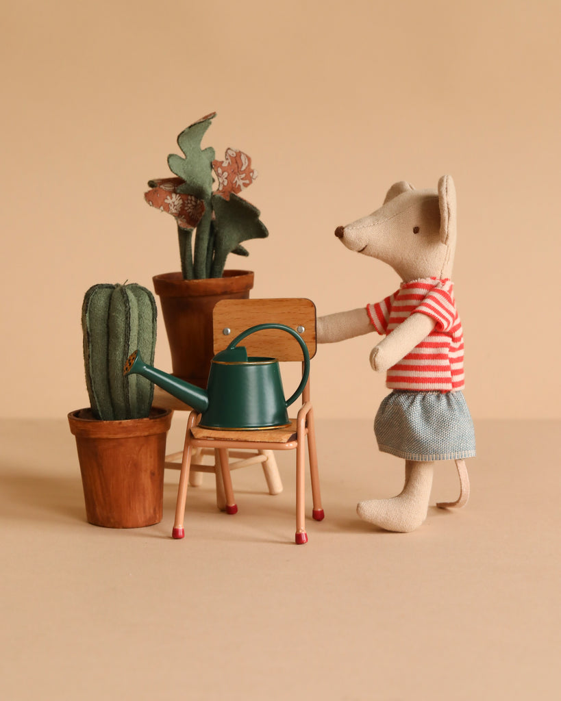 A Maileg Little Green Thumb Garden Set features a mouse in a red striped shirt and blue shorts standing by a flower pot and a green watering can on a school chair, all set against a beige background.