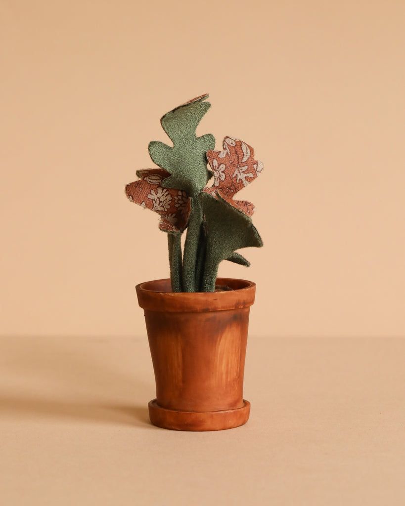 A small terracotta pot with a handmade fabric plant, featuring green and patterned leaves, complements the Maileg Dancing Mouse in Daybed – Cozy Morning Set, styled on a plain beige background.