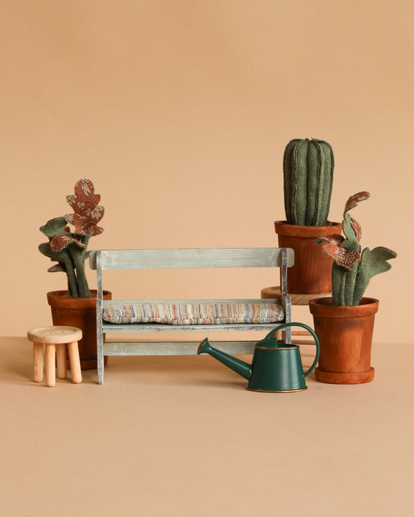 The Maileg Garden Hideaway Set, featuring a light blue bench, table & stool, potted cacti, leafy plants, and a green watering can, is displayed on a beige surface with a matching beige background.