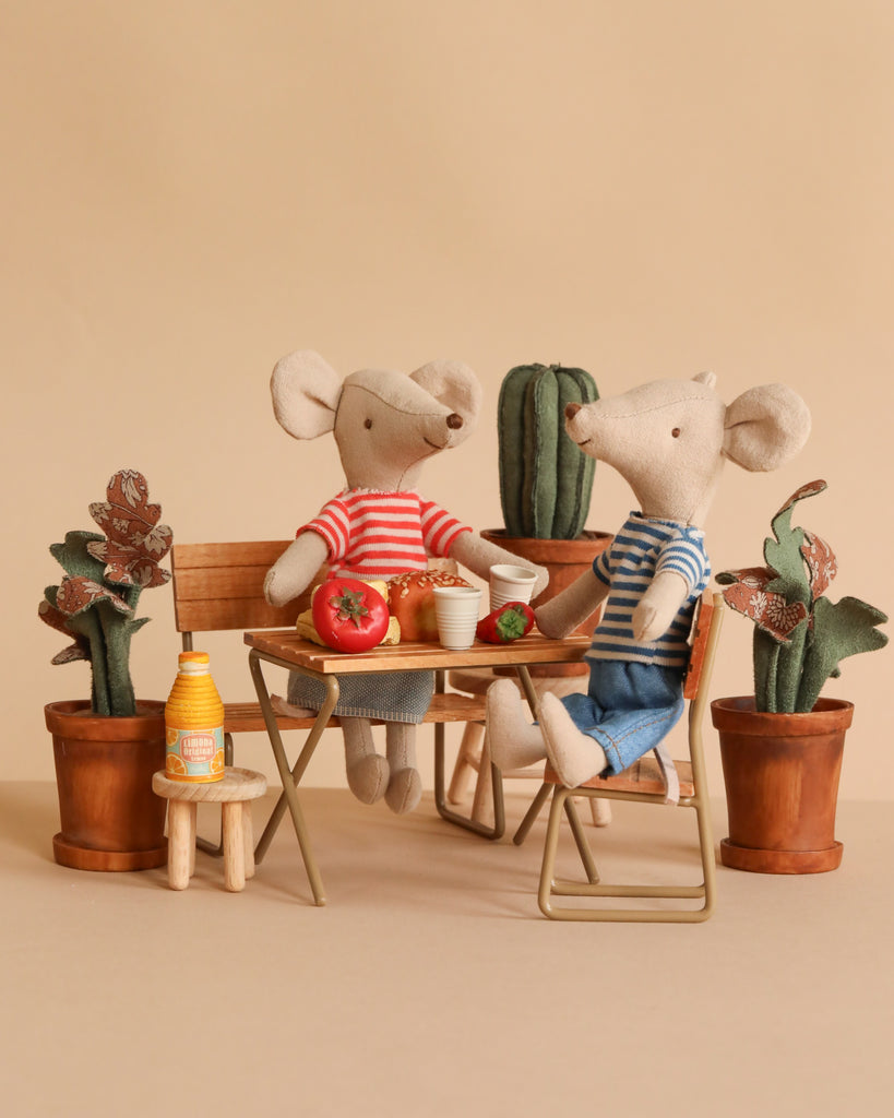 Two Maileg mice from the Picnic in the Garden Set wear red and blue striped shirts and sit at a small table with food, drinks, and potted plants, creating a cozy, playful scene.