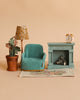 The Maileg Cozy Living Room Set features a teal armchair, floral rug, potted plant, patterned lamp, blue fireplace with logs, and a small alarm clock on the mantel, all set against a beige backdrop.