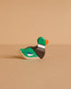 A small, handcrafted Holztiger Mallard Duck, Sitting—painted green, brown, white, orange, and blue—rests on a beige surface with a matching background, showcasing the charm and play value of HOLZTIGER figures.