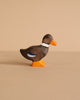 The Holztiger Duck, Standing is a small, handcrafted wooden figurine painted brown with an orange beak and feet, a white collar, and a blue accent on its wing, shown on a beige surface with a beige background.