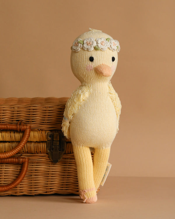 The Cuddle + Kind Duckling, a soft yellow hand-knit plush toy with a floral headband, leans against a woven wicker basket on a light brown background.