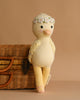 The Cuddle + Kind Duckling, a soft yellow hand-knit plush toy with a floral headband, leans against a woven wicker basket on a light brown background.