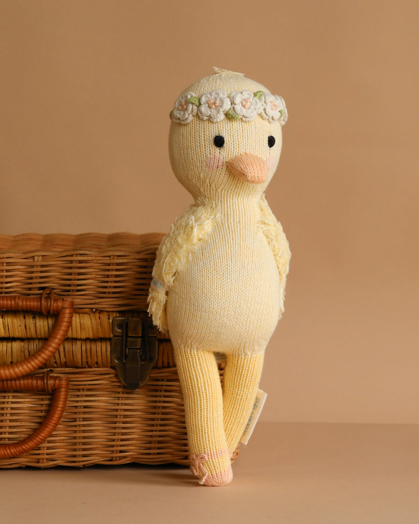 The Cuddle + Kind Duckling, a soft yellow hand-knit plush toy with a floral headband, leans against a woven wicker basket on a light brown background.