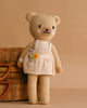 Cuddle + Kind Goldie The Honey Bear is a hand-knit teddy bear with a beige body and a cream apron dress adorned with a small yellow flower, pictured in front of a wicker basket on a plain tan background.